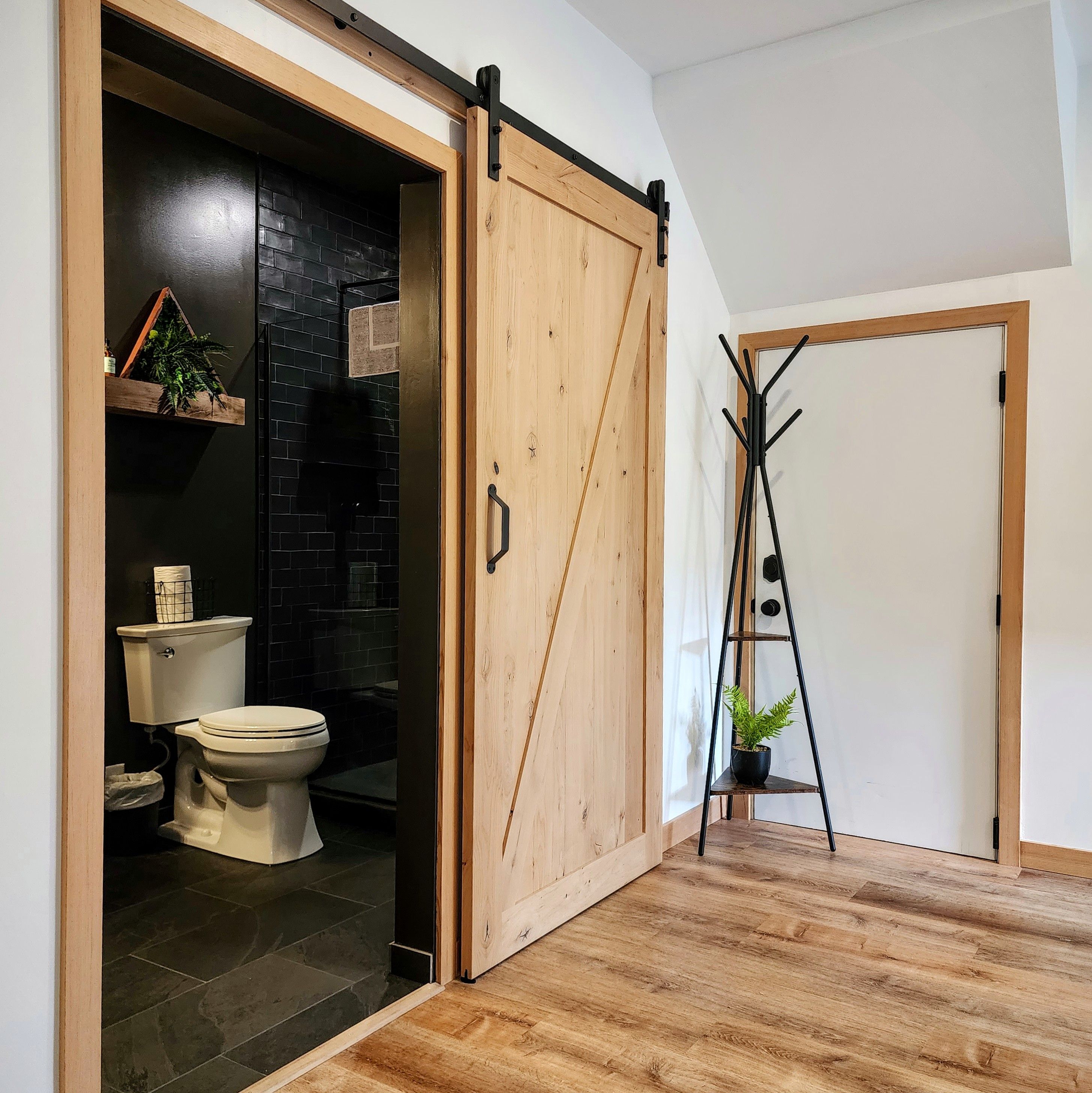 Converted garage suite in Ucluelet: a wood barn door open to a dark-tiled bathroom, vinyl plank floor and fir trim.